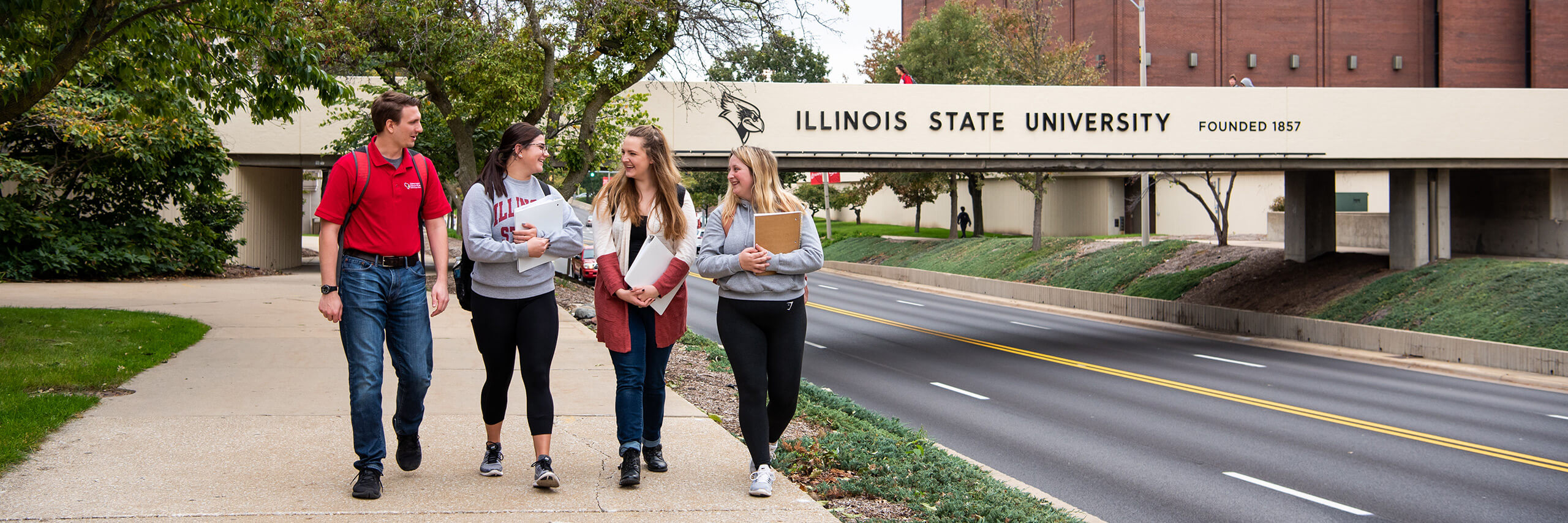 Four Illinois State students walk path in front of College Avenue bridge.
